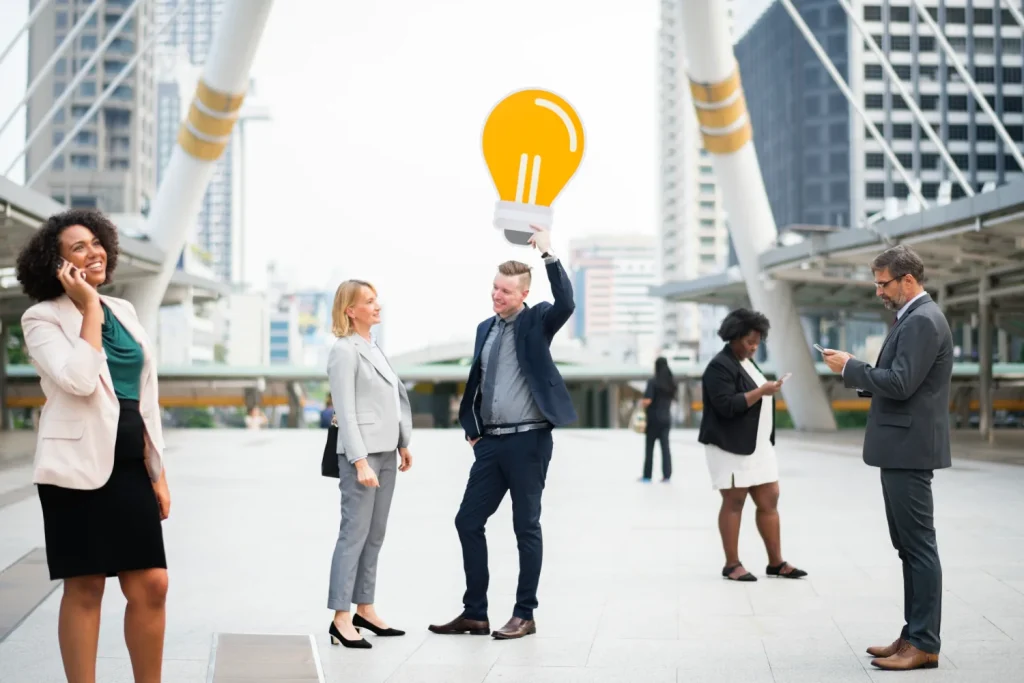 Business professionals networking outdoors, with one person holding a large yellow lightbulb cutout.