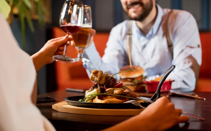 A delicious plate of grilled skewers served at a restaurant table with two people toasting with drinks.