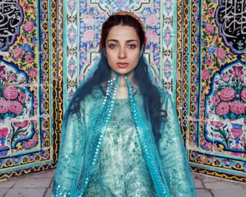 Persian woman in traditional turquoise attire with intricate embroidery, standing against vibrant floral tilework.