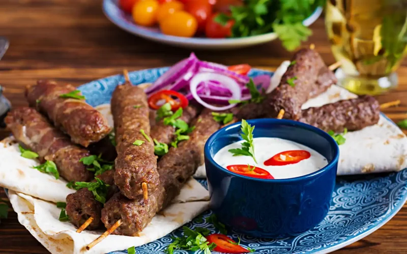 A plate of Persian koobideh kebabs served with yogurt sauce, onions, and fresh herbs.